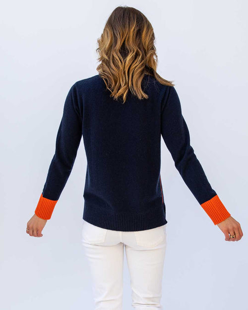 Woman wearing navy cashmere sweater with orange cuffs and white pants, viewed from the back against a plain background