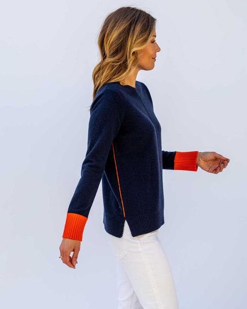 LIFESTYLE / EDITORIAL: Woman in navy cashmere sweater with orange cuffs and white pants, side profile against plain