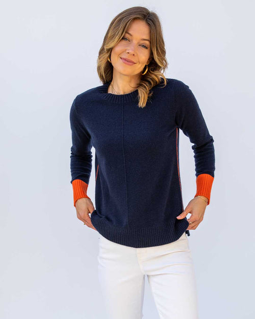 Woman wearing navy cashmere sweater with orange cuffs and white pants against a plain light background