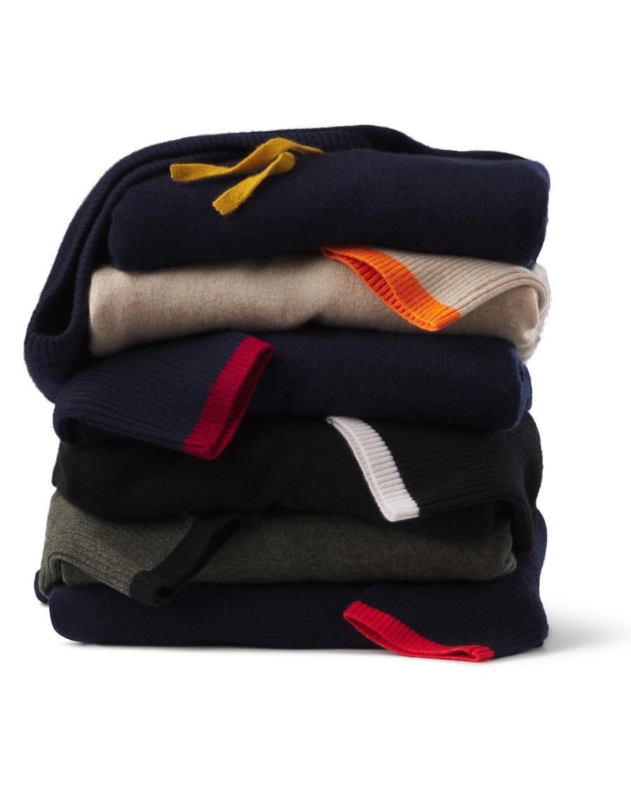 Six folded cashmere pullovers and hoodies in a stack, each showing its second color as a knitted drawstring or cuff edge, on white.